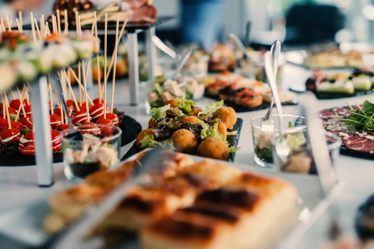 A spread of assorted appetizers on a buffet table, including skewers, salads, pastries, and various bite-sized snacks, with tongs and serving utensils visible. Foods that were once luxury items.