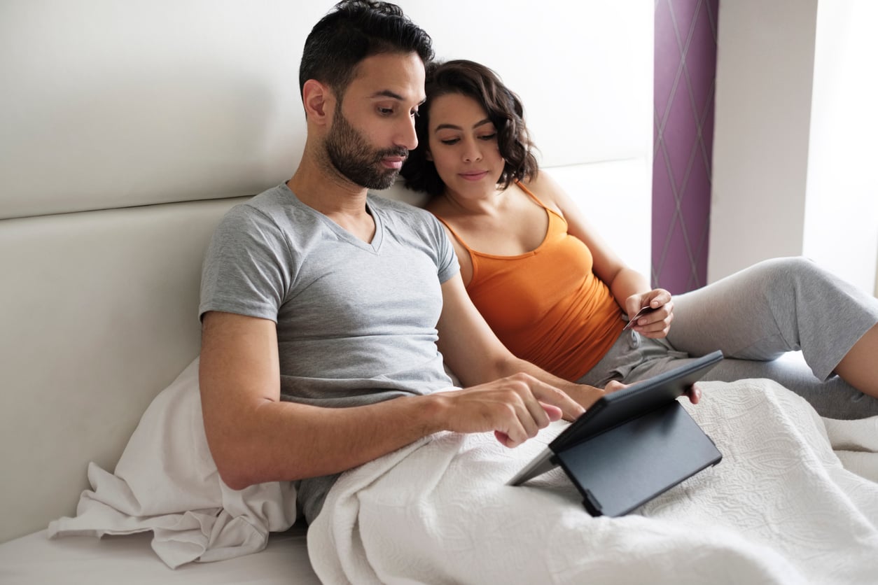 A man and woman sit together in bed under a white blanket, both looking at a tablet. The man wears a gray t-shirt, and the woman wears an orange tank top and gray pants. They appear relaxed and engaged with the device.