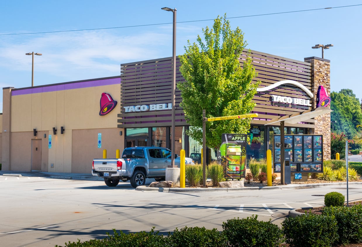 A silver pickup truck is parked at the drive-thru of a Taco Bell restaurant on a sunny day. The Taco Bell sign and menu board are visible, and a tree stands near the building.