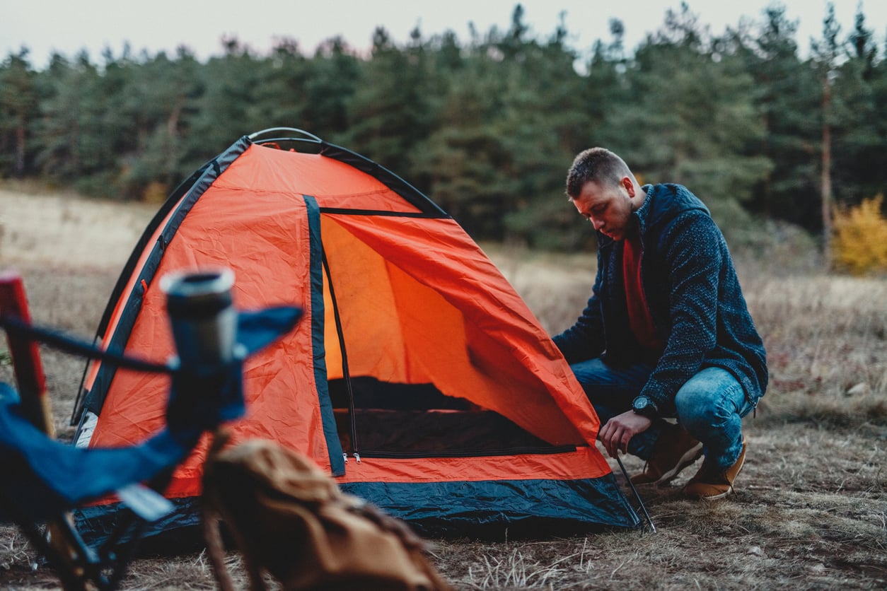 A man kneels next to an open orange tent in a grassy field near a forest, preparing his campsite. Camping gear is visible in the foreground, showcasing one of the best budget-friendly hobbies for outdoor enthusiasts.
