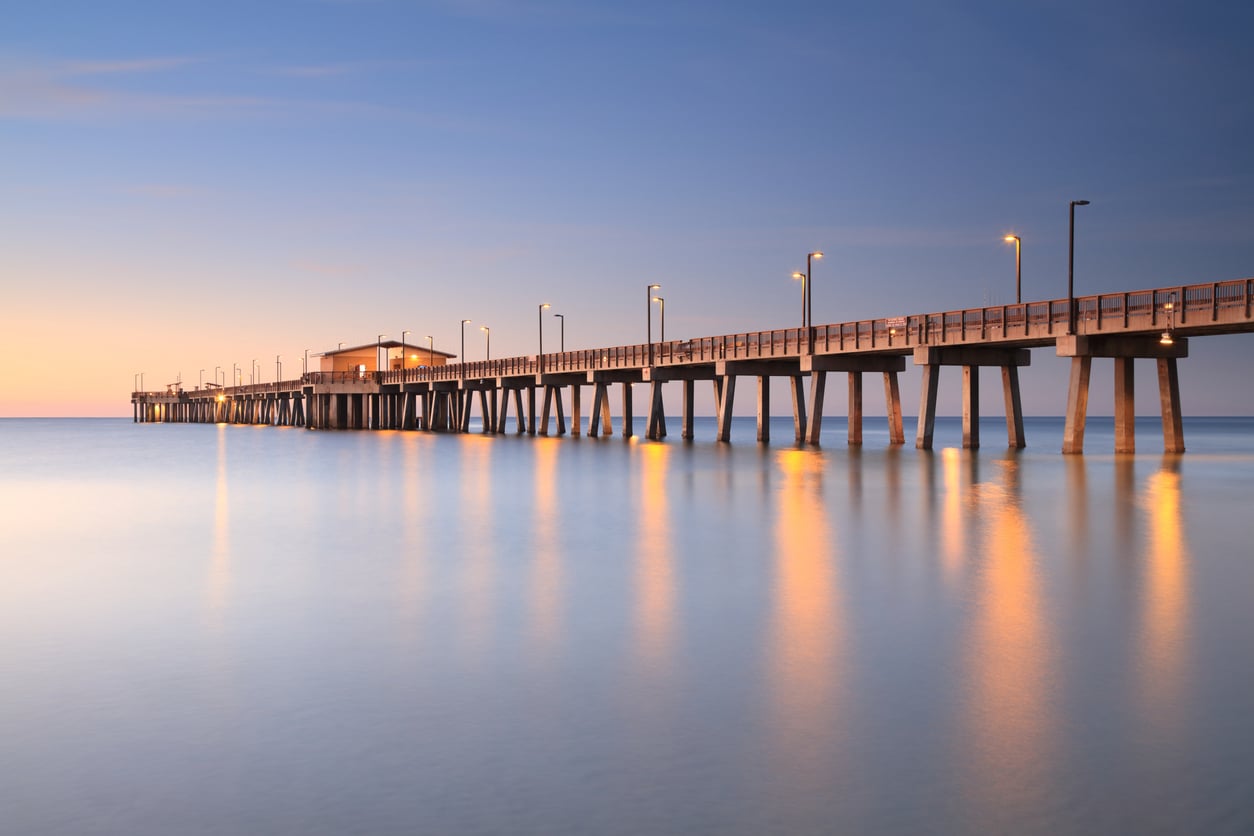 A long wooden pier with railings and lights extends over calm water at sunset, reflecting warm light. The sky is clear with soft hues of blue and orange, and a small building sits at the end of the pier.