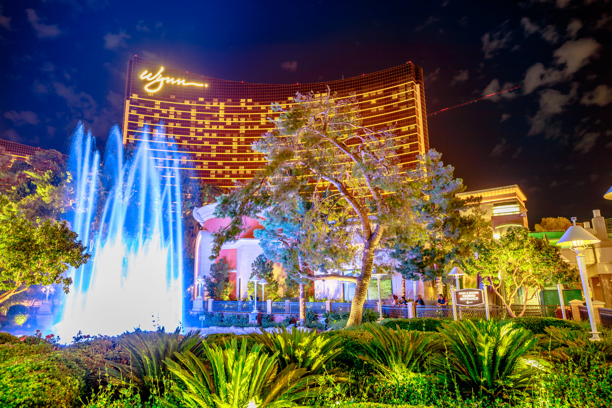 A brightly lit fountain and lush greenery in front of the illuminated Wynn hotel at night in Las Vegas, with glowing signage atop the curved building—perfect for capturing memories while exploring Vegas money-saving tips.