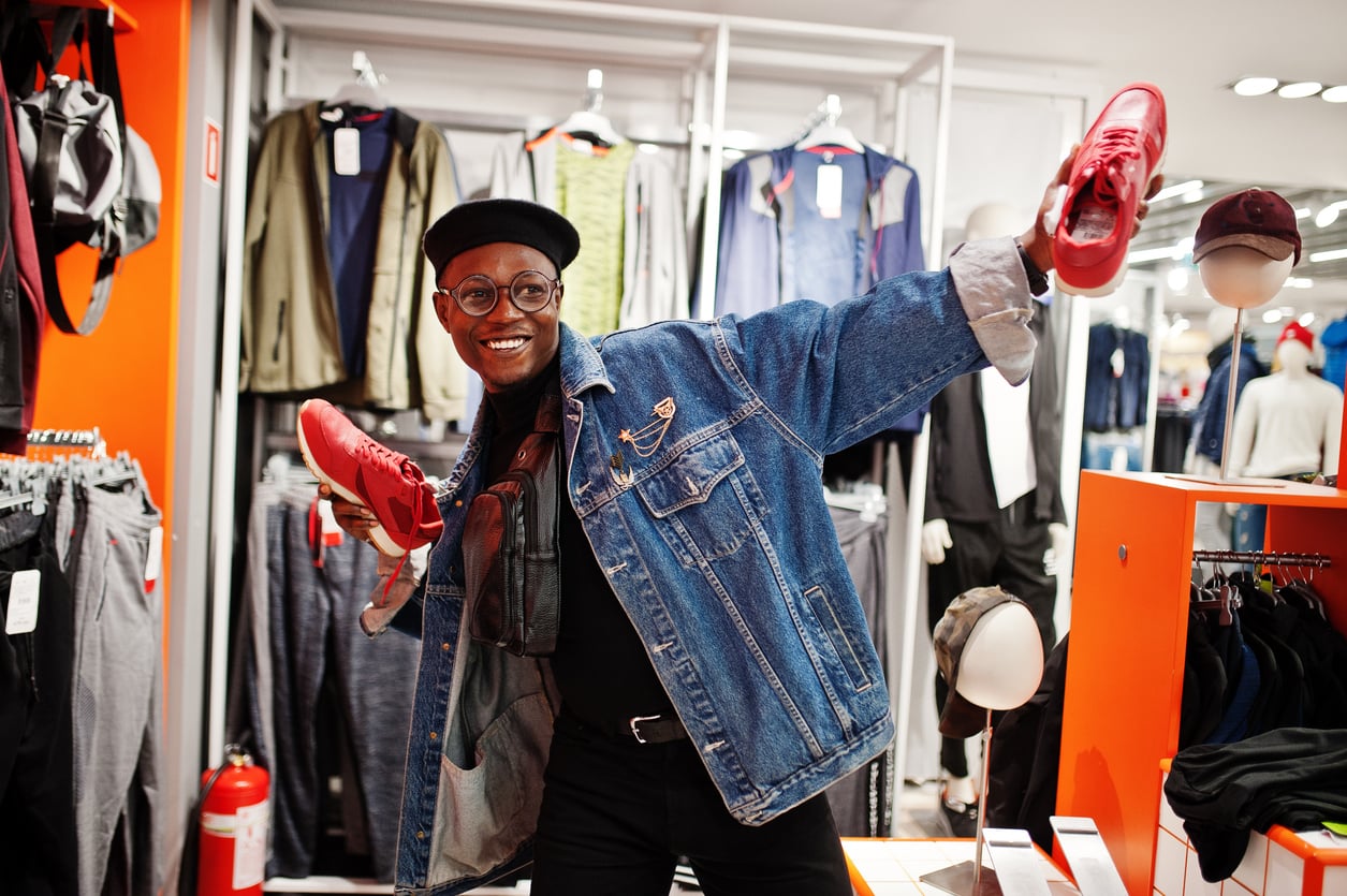 A smiling person in a denim jacket and cap playfully poses in a clothing store, holding up a pair of red sneakers—one of those products men skip buying—surrounded by racks of clothes and accessories.