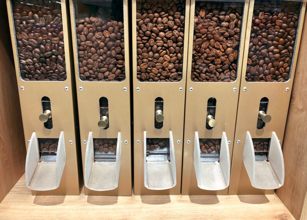 Five transparent dispensers filled with different types of coffee beans, each with a metal lever and a silver chute for dispensing the beans into containers below. The dispensers are set in a wooden frame.