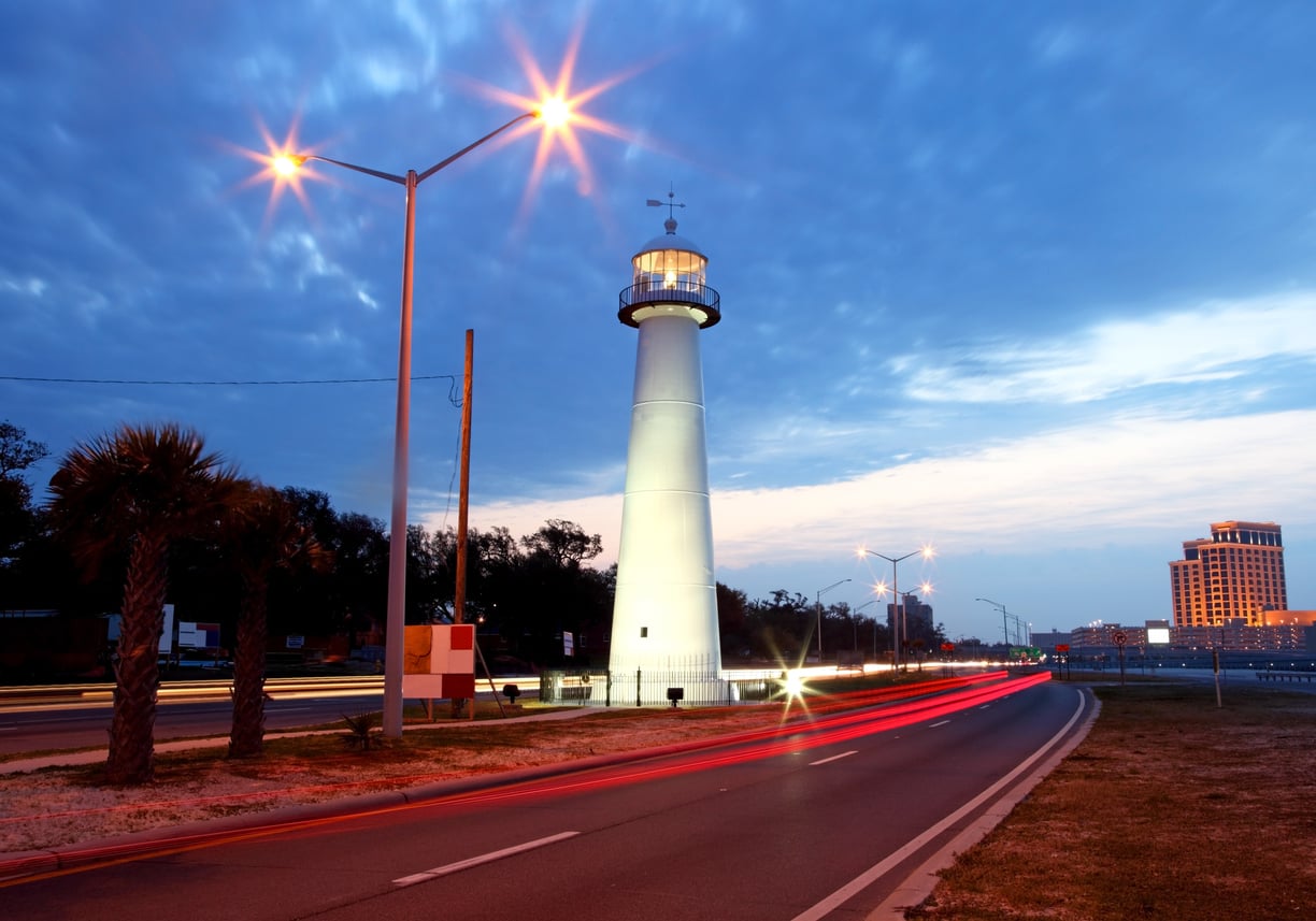 A lit lighthouse stands beside a road at dusk, with car light trails curving past. Palm trees and streetlights line the road, while a building rises in the background under a cloudy blue sky. Cheaper resort towns.