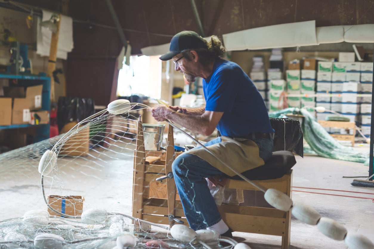 A man wearing a blue shirt, cap, and glasses repairs a fishing net inside his workshop—an example of Small Business States—surrounded by boxes, shelves, and other nets. Seated on a wooden bench, he is fully focused on his craft.