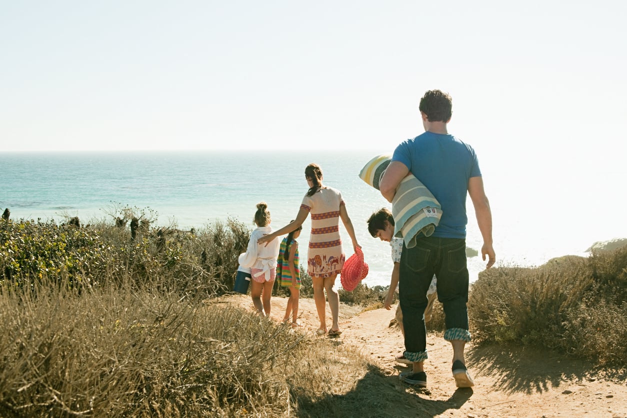 A family walks on a dirt path toward the beach, embracing moments that support mental health. An adult carries a child, while others walk ahead holding hands. The ocean and blue sky are visible beyond dry grass and bushes.