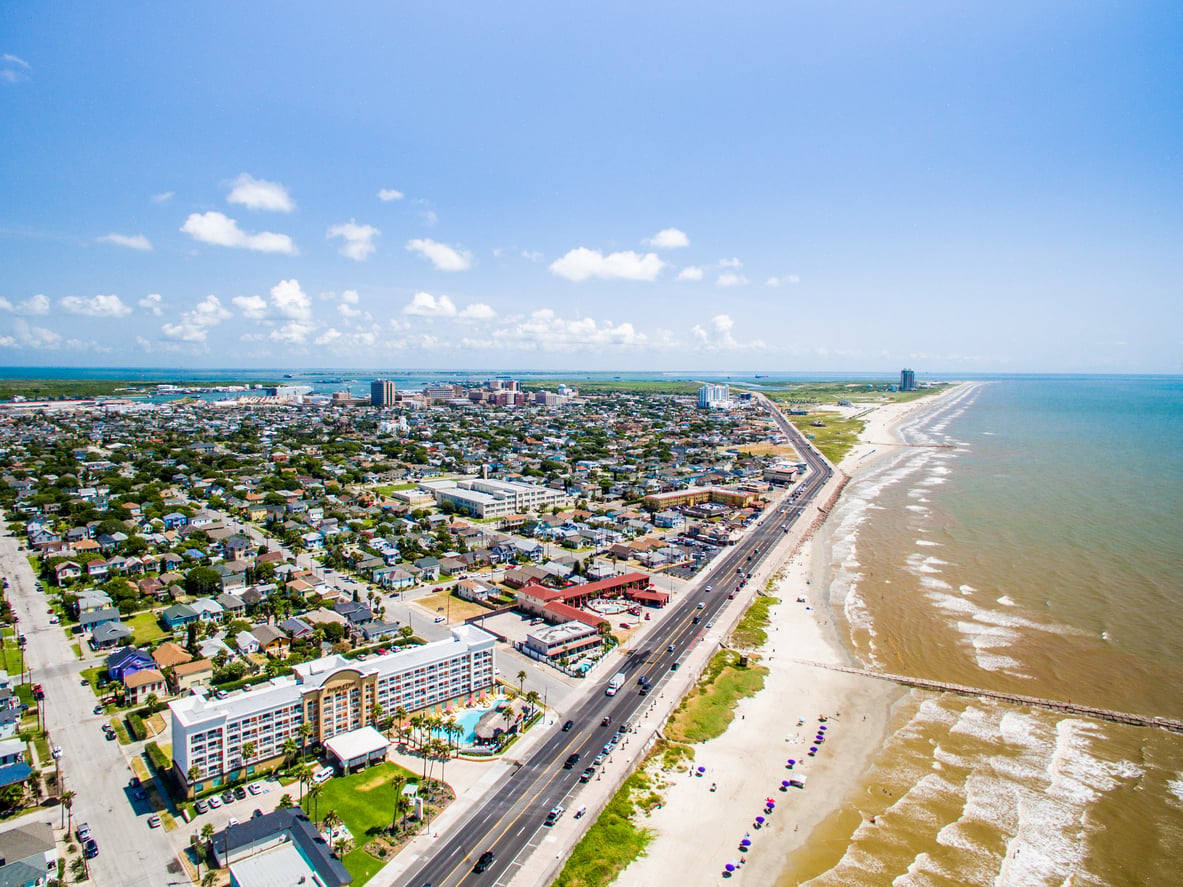 Aerial view of a coastal city with a road running alongside a sandy beach, buildings and houses on one side, and the ocean stretching into the distance under a clear blue sky.