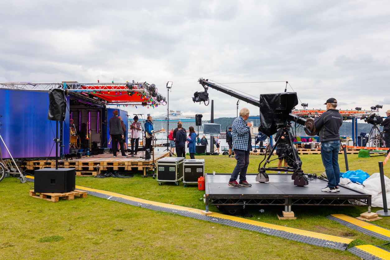 A TV crew films a live outdoor broadcast on a grassy field. Large cameras, lights, and crew members are visible, with people preparing on stage—perhaps sharing concert ticket hacks—with water and buildings in the background.