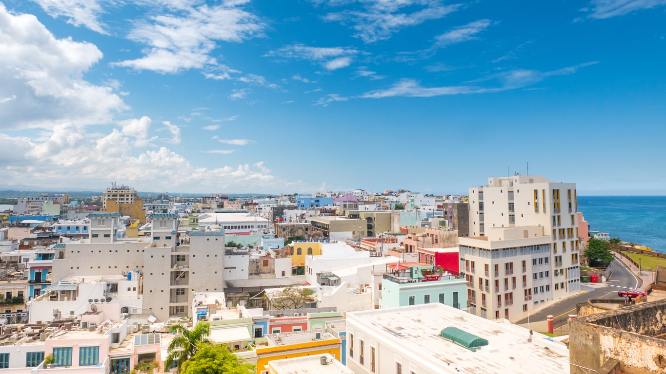 A scenic view of a coastal city with colorful buildings under a partly cloudy blue sky, with the ocean visible on the right side.