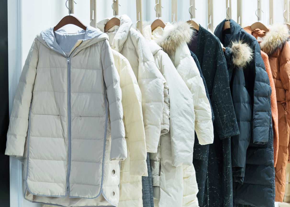 A row of winter clothes, including puffer jackets and parkas in white, cream, black, and orange, hang neatly on wooden hangers in a bright store.