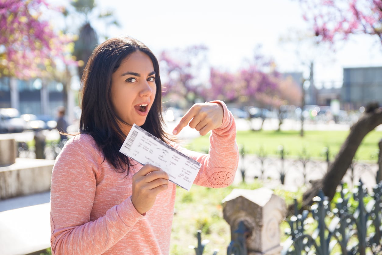 A woman in a pink sweater stands outside in a park, smiling with her mouth open and pointing excitedly at a ticket she is holding—she’s just scored big using concert ticket hacks. Pink-blossomed trees bloom in the background.