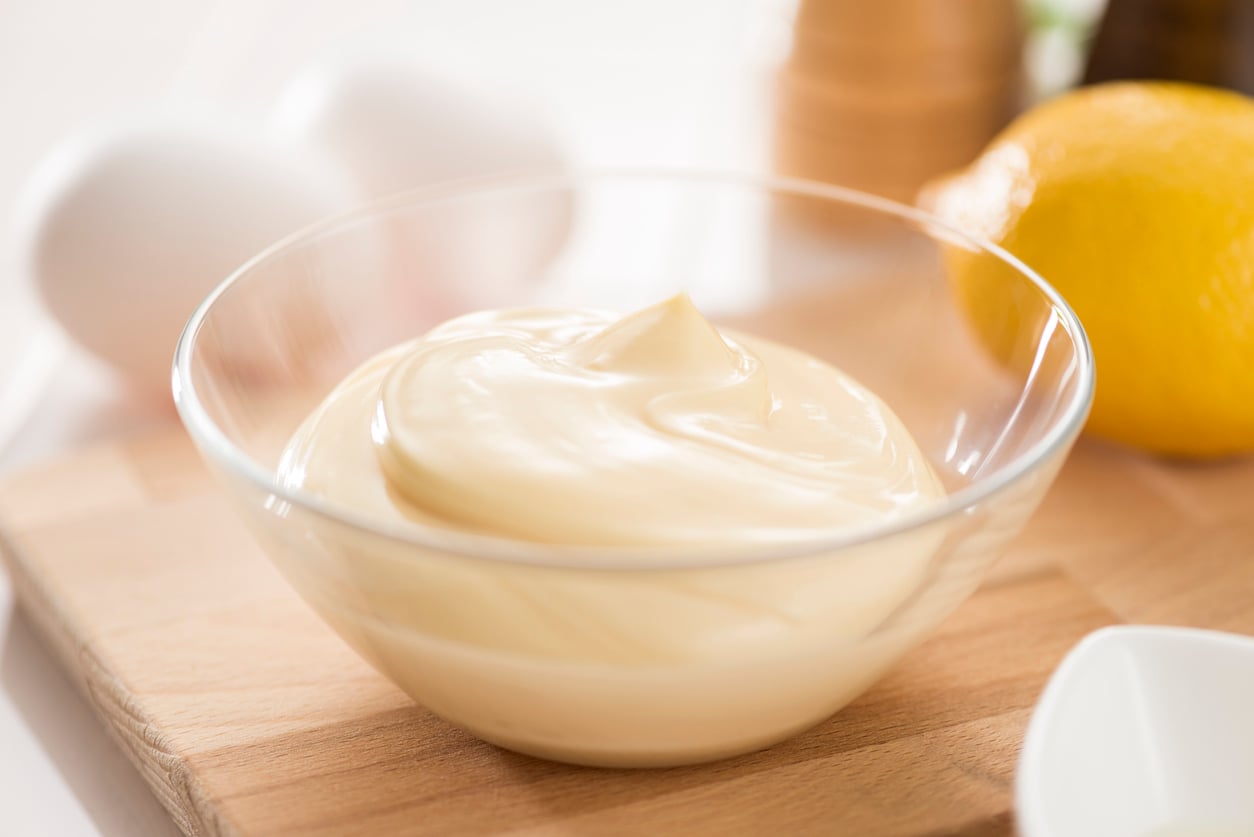A clear glass bowl filled with creamy mayonnaise sits on a wooden board. In the background, there are two eggs, a lemon, and blurred containers—perfect for exploring recipes with egg substitutes.