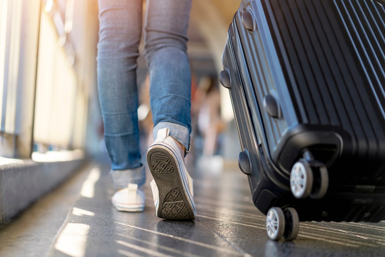 A person wearing jeans and sneakers walks while pulling a black rolling suitcase, viewed from behind, in a bright indoor setting, possibly an airport or station.