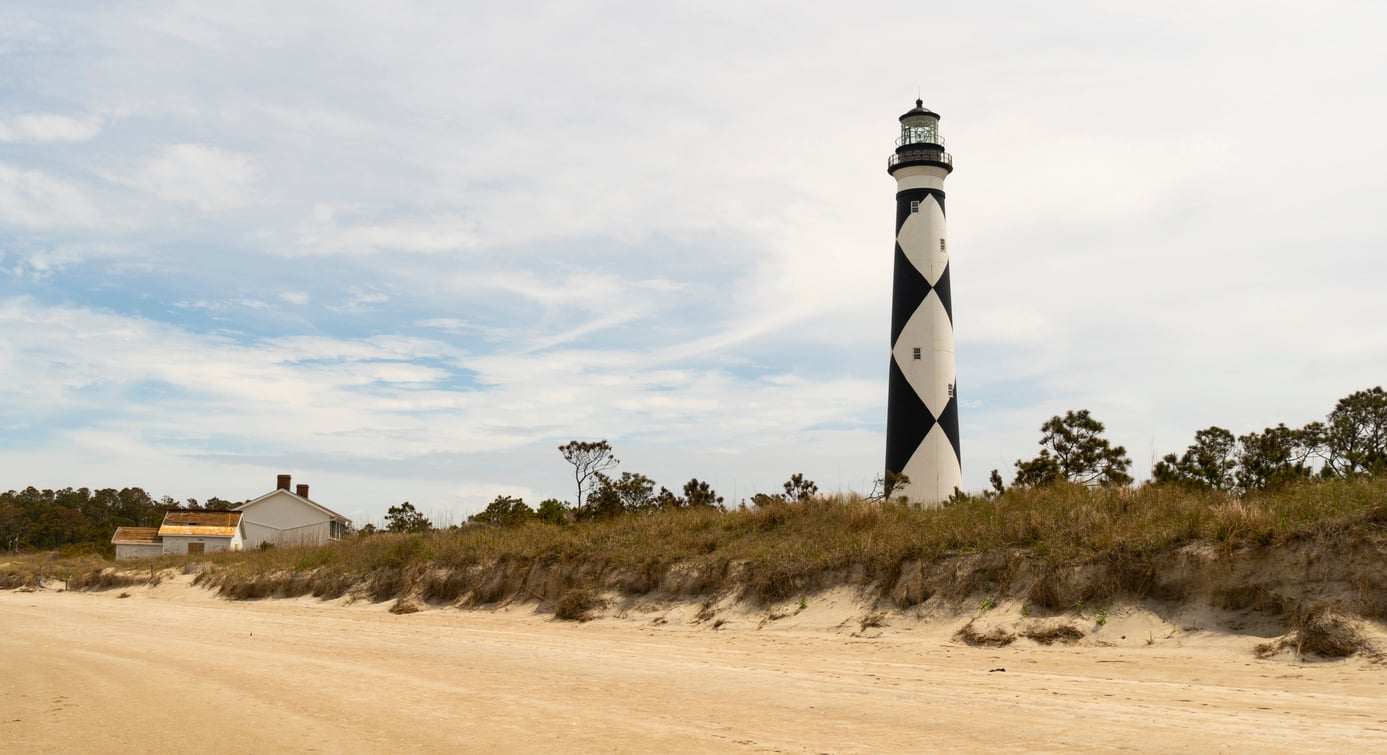 A tall lighthouse with a black and white diamond pattern stands near a sandy beach, with grassy dunes and a few buildings and trees in the background under a partly cloudy sky.