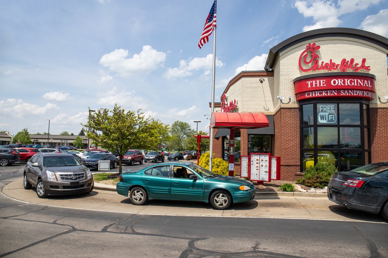 Cars line up at a Chick-fil-A drive-thru on a sunny day, with an American flag flying above the restaurant and several vehicles waiting in two lanes. A sign on the building advertises free WiFi and original chicken sandwiches.