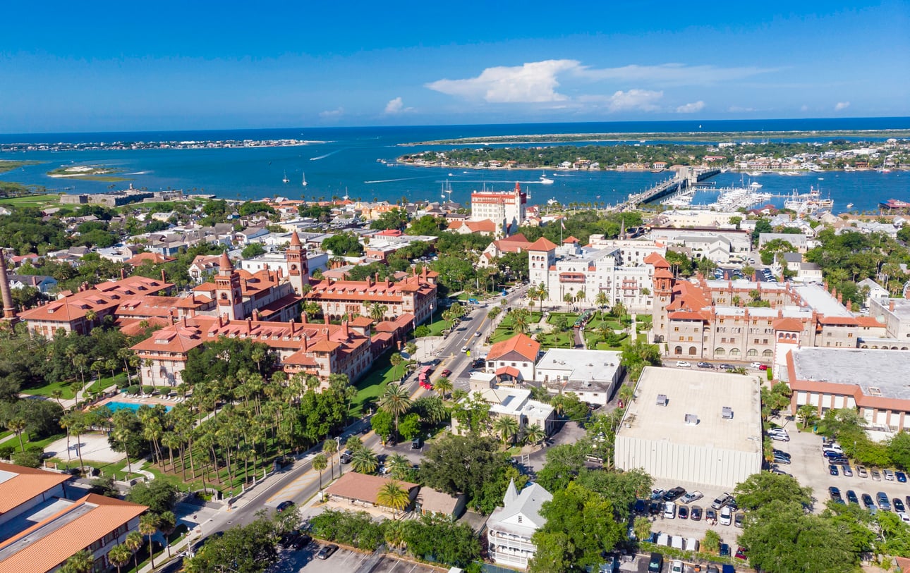 Aerial view of a coastal city with historic red-roofed buildings, lush greenery, a bridge over blue water, and boats in the harbor under a clear blue sky. Road trip food destinations. 