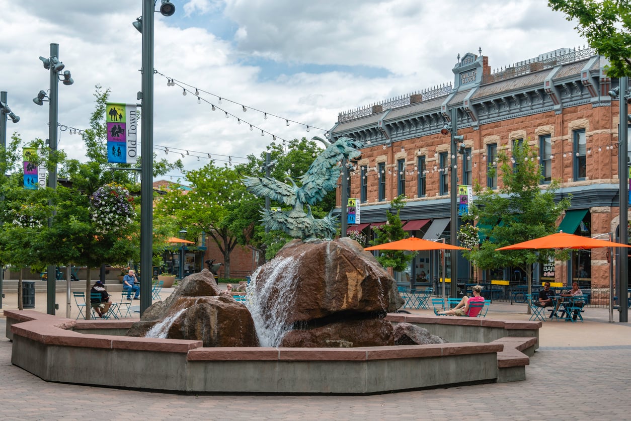 A plaza in one of the nation’s affordable college towns features a rock fountain, outdoor seating with orange umbrellas, string lights, and a bronze sculpture. Historic brick buildings and shops stand in the background under a cloudy sky.