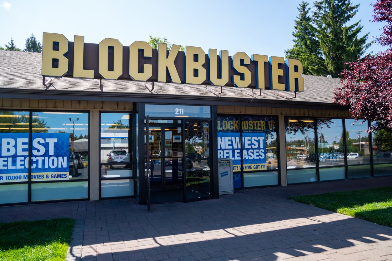 A Blockbuster store with large yellow sign, glass doors, and windows displaying posters that read “Best Selection,” “Newest Releases,” and “1991 Prices.” Trees and blue sky are visible in the background.