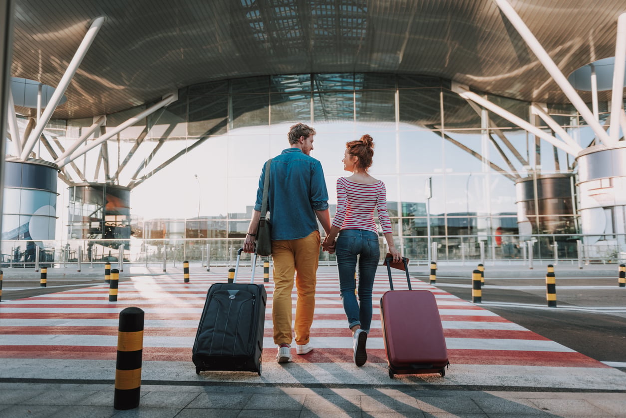 A couple holding hands and pulling suitcases walk across a crosswalk toward a modern glass airport terminal, seen from behind on a sunny day.