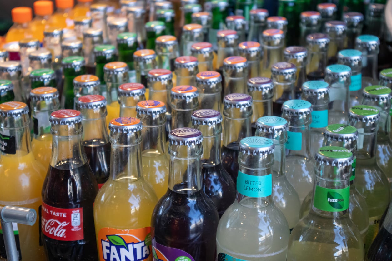 Rows of assorted glass bottles containing various soft drinks, including Fanta, Coca-Cola, and other colorful beverages with different labels and caps, showcase a tempting array of often overpriced products arranged closely together.