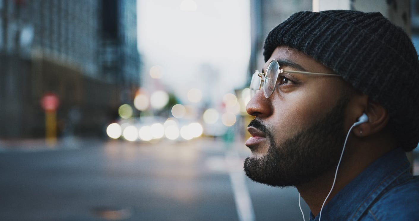 A man wearing glasses, a knit beanie, and earphones—perhaps pondering the products men skip buying—looks thoughtfully into the distance on a city street at dusk, with blurred lights and buildings in the background.
