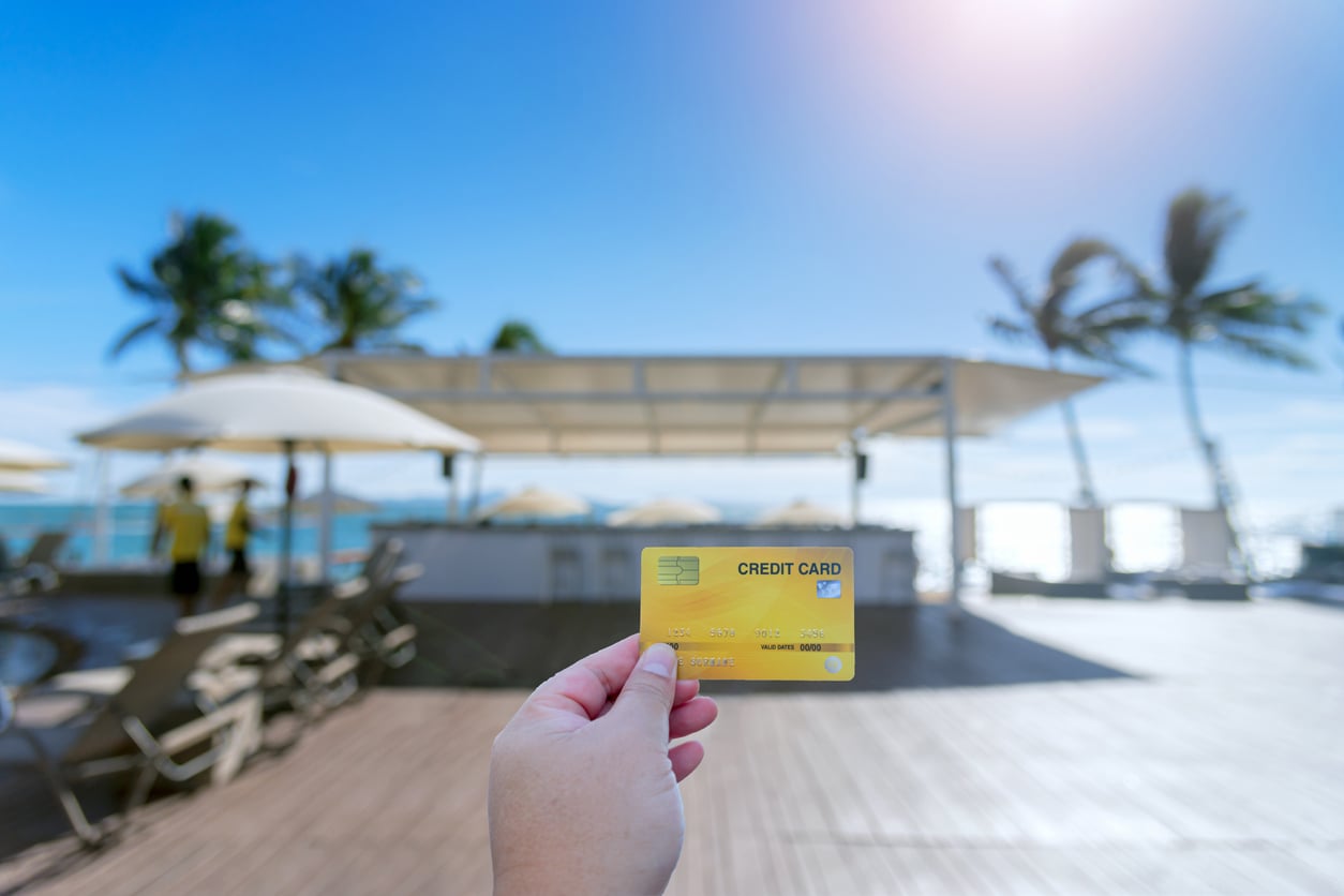 A hand holds a yellow credit card—one of the best travel hacks—in front of a sunny beachside bar with palm trees, lounge chairs, and umbrellas. The background is slightly blurred, highlighting a bright blue sky.