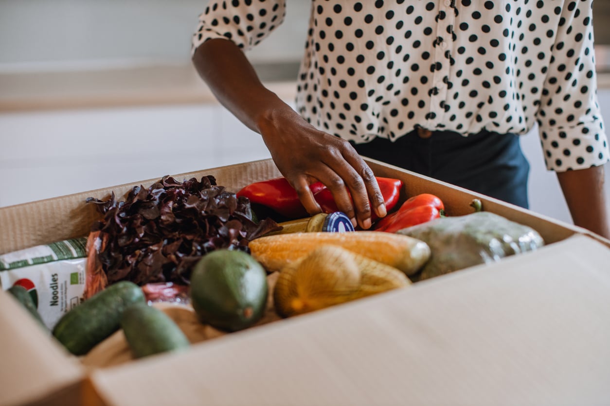 A person in a polka dot shirt reaching into a cardboard box filled with fresh vegetables, including avocados, lettuce, zucchini, and red peppers.