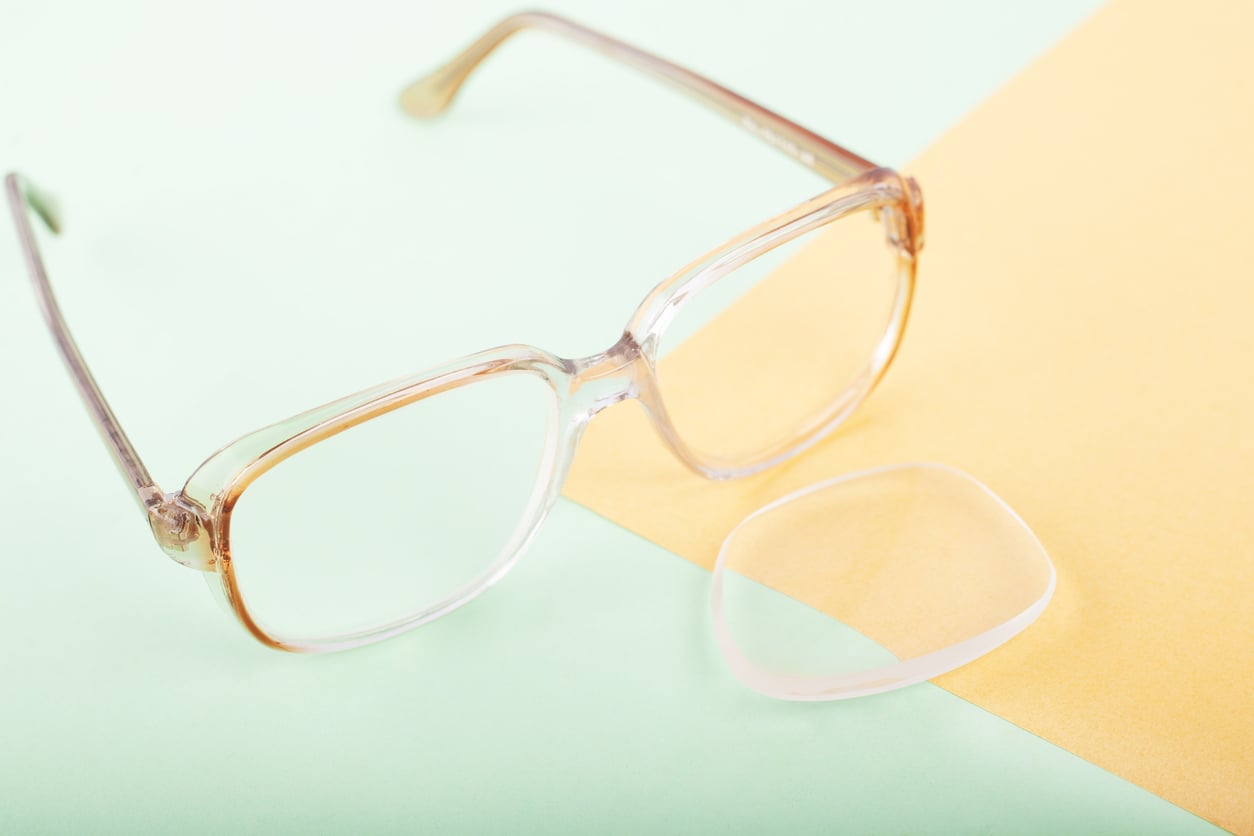 A pair of transparent eyeglasses with one detached lens lying next to the frame on a pastel green and yellow background, subtly reflecting the consequences of delayed purchases.
