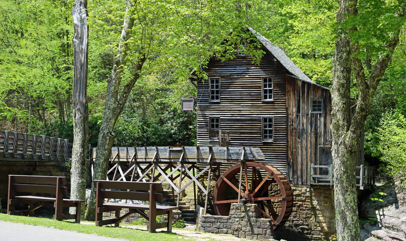 A rustic wooden watermill with a large water wheel sits among green trees next to a path, with benches nearby and sunlight filtering through the leaves.