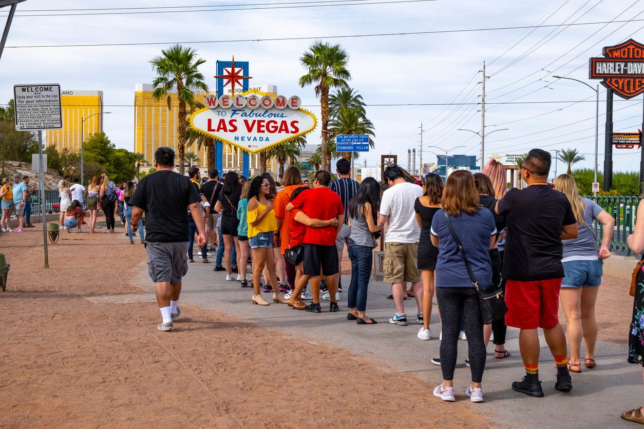 A long line of people waits to take photos in front of the famous "Welcome to Fabulous Las Vegas, Nevada" sign, one of the top Las Vegas attractions, on a sunny day with palm trees and buildings in the background.