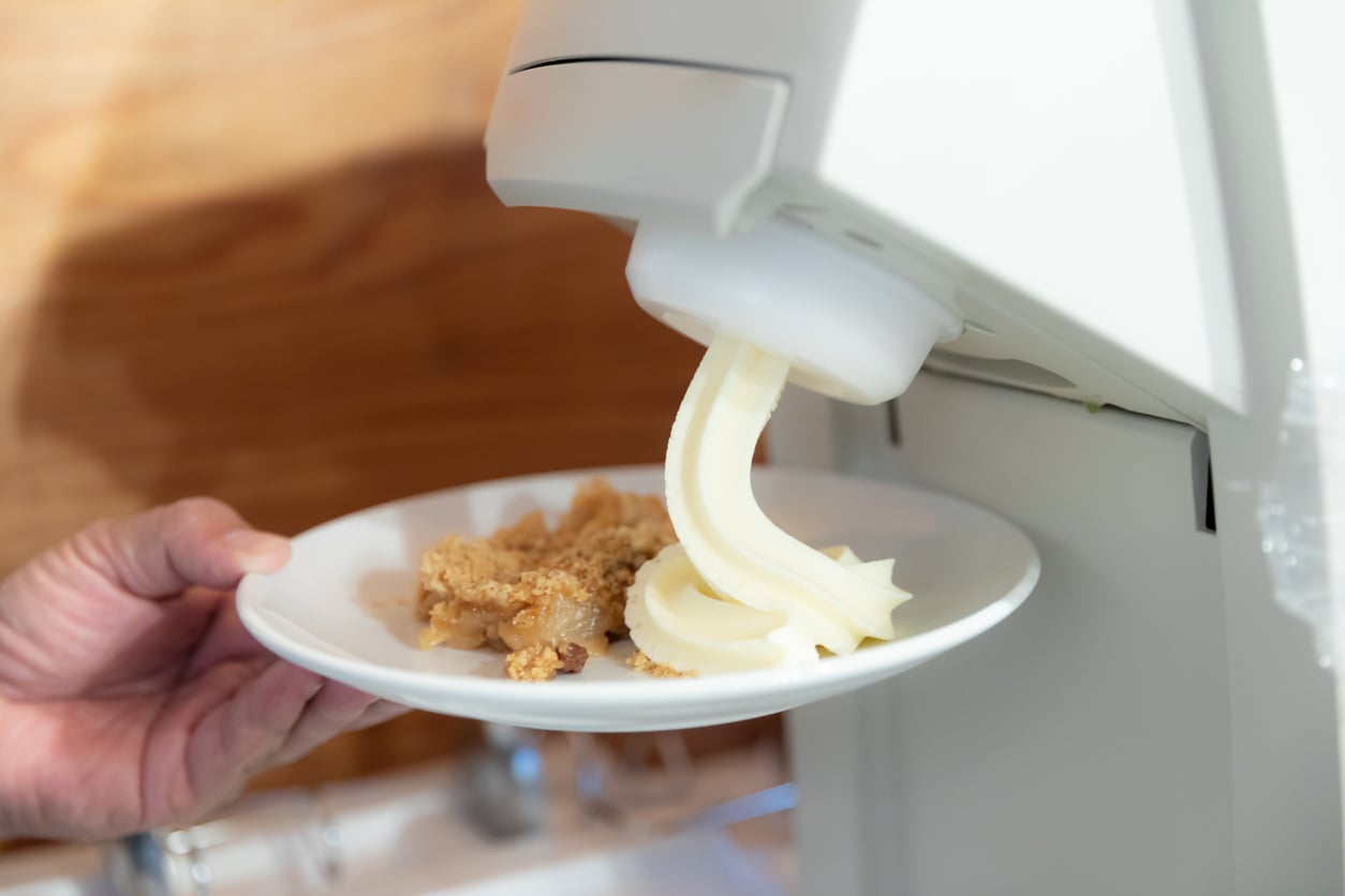 A hand holds a plate under a soft-serve ice cream machine, dispensing vanilla ice cream next to a serving of crumble dessert.