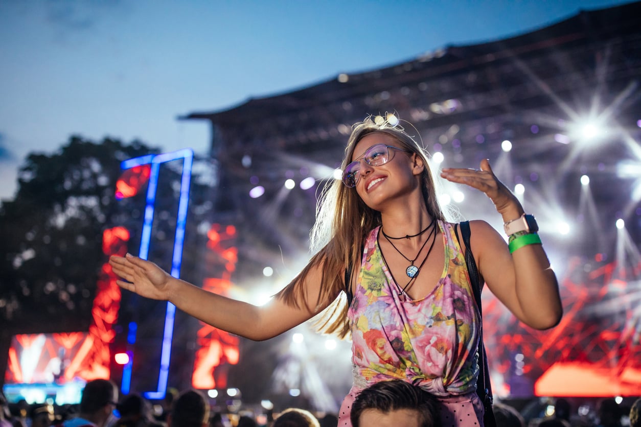 A young woman wearing glasses and a floral top smiles and dances on someone’s shoulders at an outdoor music festival, colorful stage lights shining behind her—enjoying the moment thanks to smart concert ticket hacks.