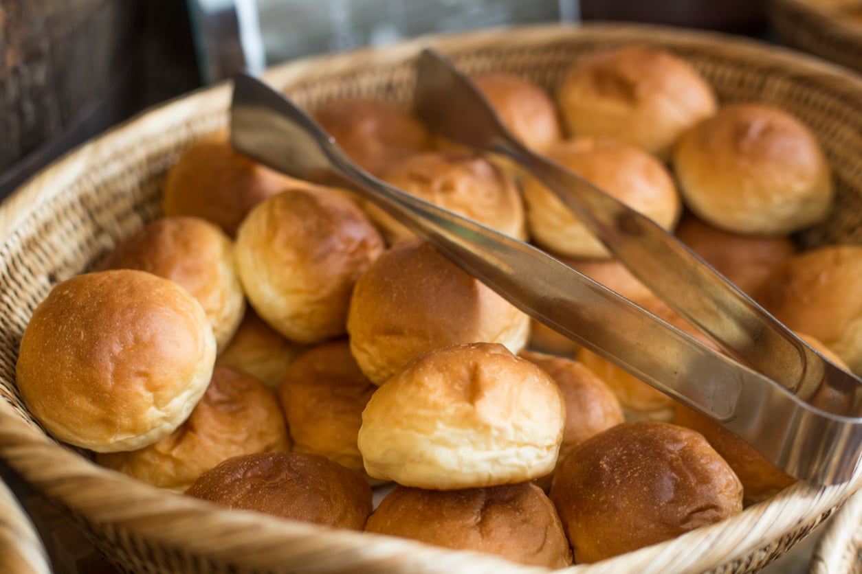 A wicker basket filled with round, golden brown bread rolls. Metal tongs rest on top of the rolls, ready for serving.