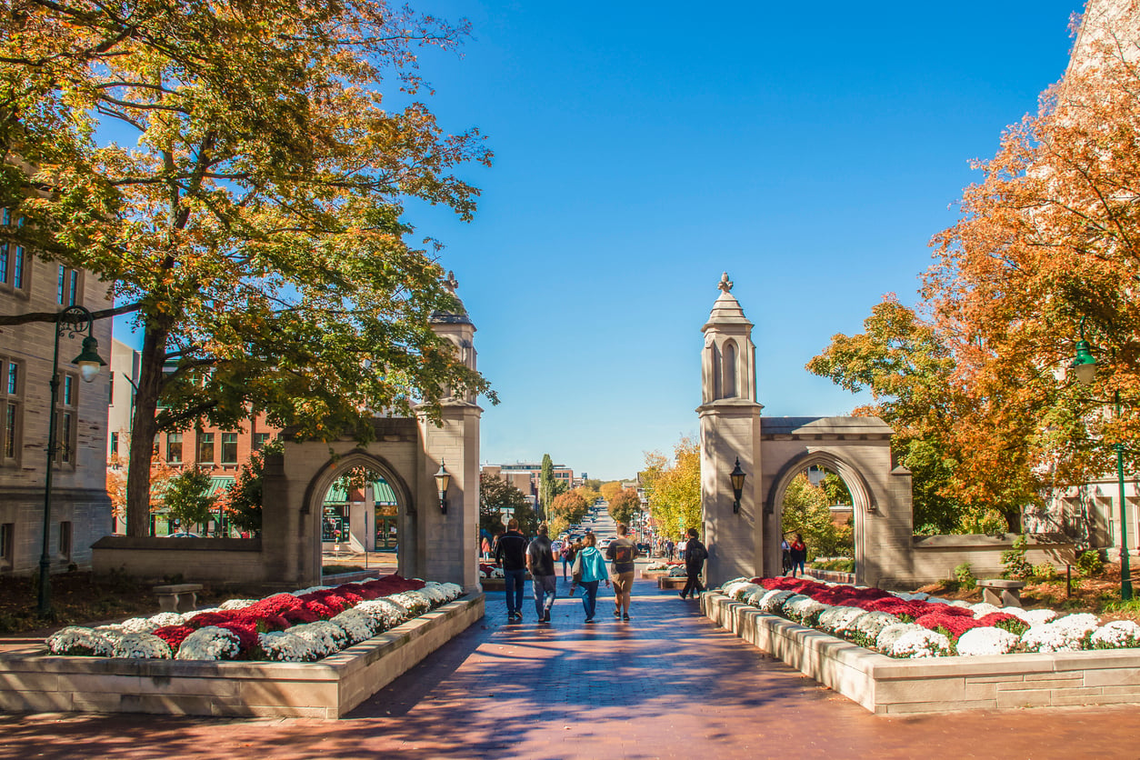 People walk through an arched stone entrance surrounded by flowerbeds and autumn trees on a bright sunny day, showcasing the charm of one of America’s affordable college towns, with campus buildings visible in the background.