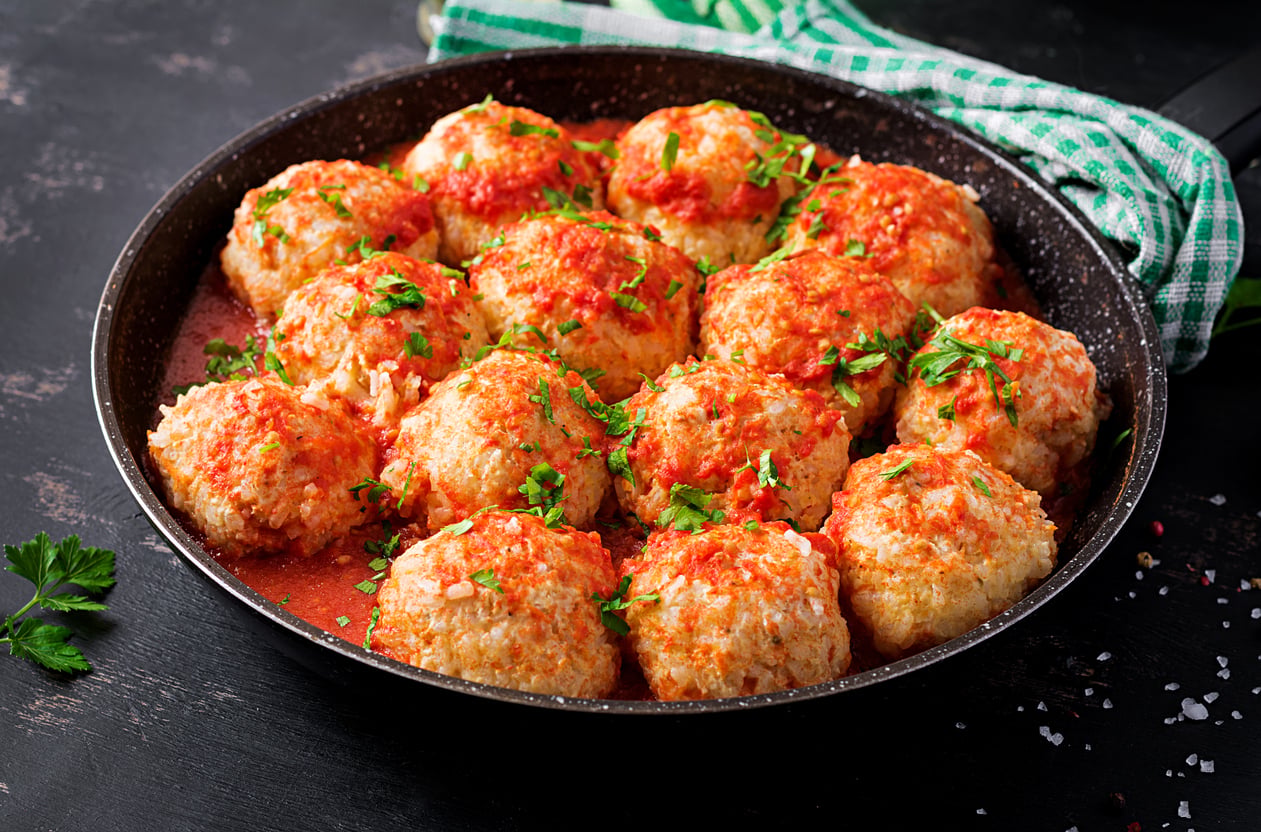 A skillet filled with golden-brown meatballs made with ground beef alternatives, topped with tomato sauce and chopped parsley, sits on a dark surface with a green-checked cloth nearby.