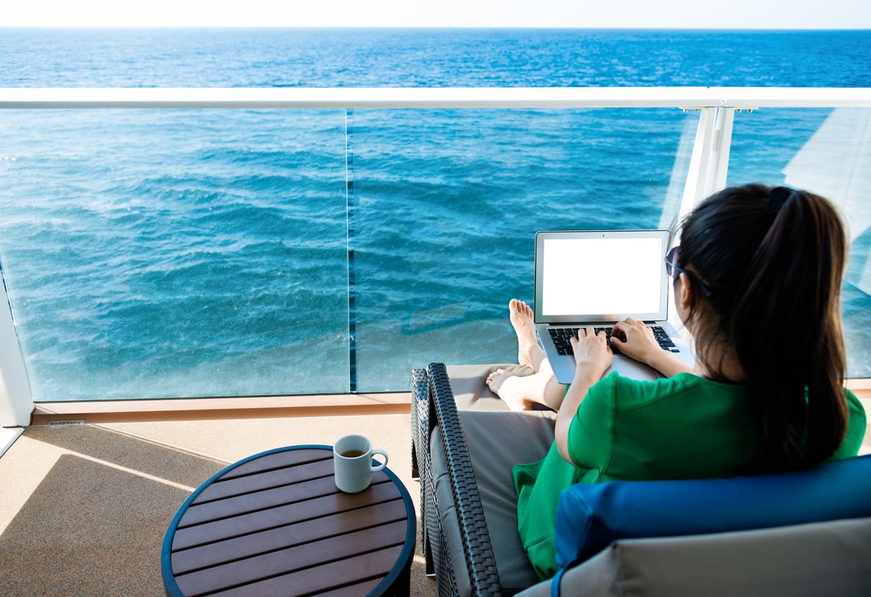 A person in a green dress works on a laptop while sitting on a lounge chair on a balcony overlooking the ocean, enjoying some travel hacks. A small round table beside them holds a white mug.