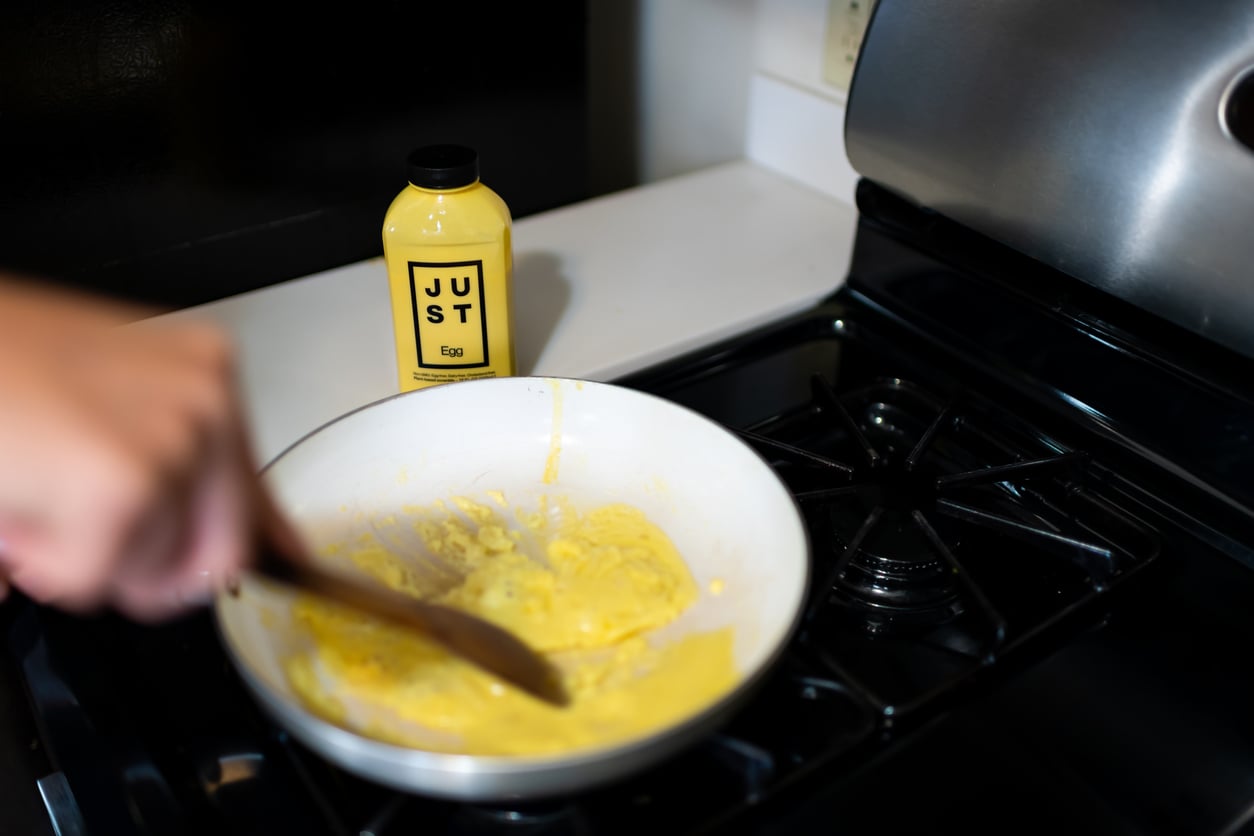 A person stirs yellow scrambled Just Egg, a popular egg substitute, in a white pan on a stove; a bottle of Just Egg is visible on the counter in the background.