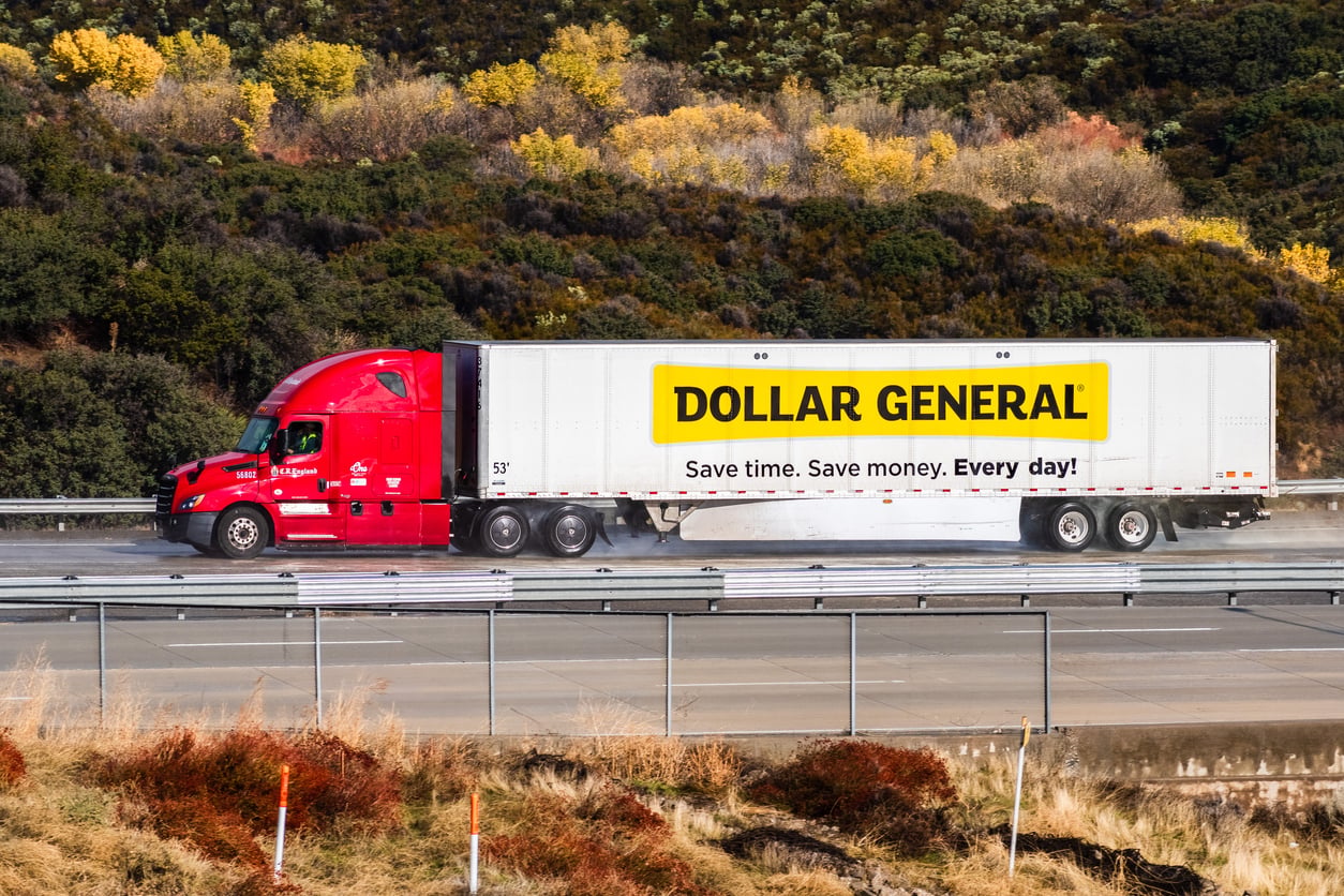 A red semi-truck with a white Dollar General trailer drives on a highway, surrounded by grassy hills and autumn trees. The trailer features the Dollar General logo and the slogan "Save time. Save money. Every day! Dollar General expansion.