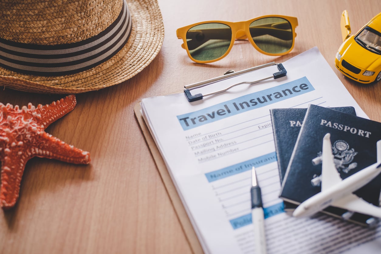 A travel insurance form on a clipboard sits on a desk, surrounded by a sun hat, sunglasses, toy car, starfish, pen, two passports, and a small airplane model—essential items for travel hacks when preparing for an overseas trip.