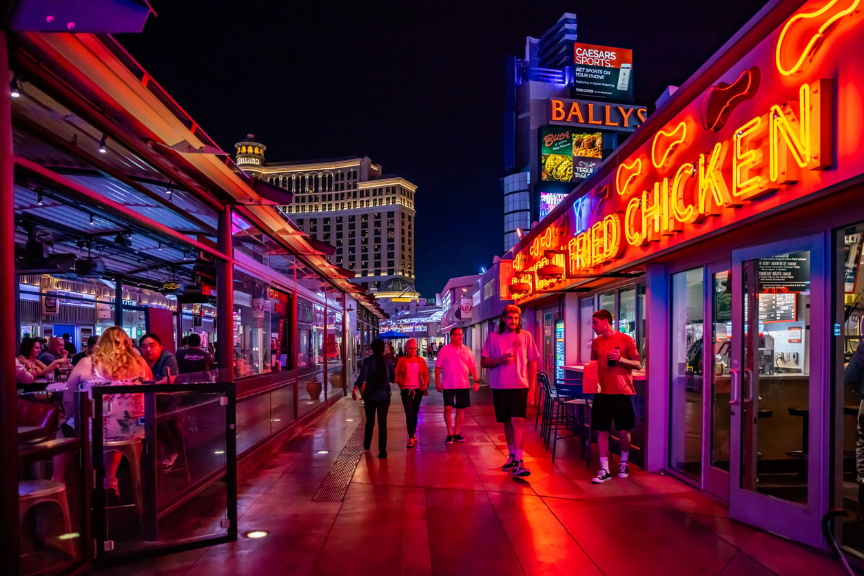 People walk along a brightly lit street at night, lined with neon signs including one for fried chicken, with restaurants and glass walls. In the distance, Bally’s and Caesars Palace hotels tower—perfect spots to try out some Vegas money-saving tips.