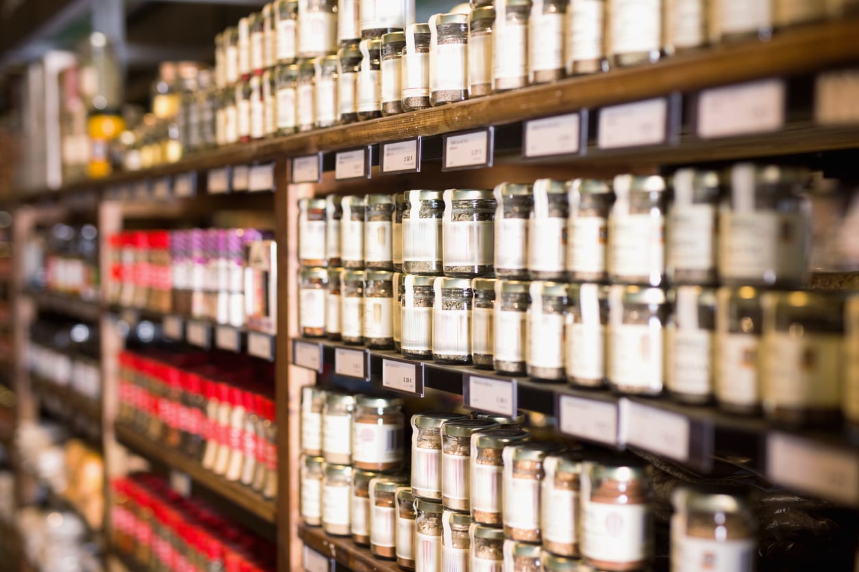 Shelves in a grocery store or market displaying rows of glass jars filled with various preserves or condiments—even some overpriced products—all neatly organized and labeled, with select jars in focus and others blurred in the background.