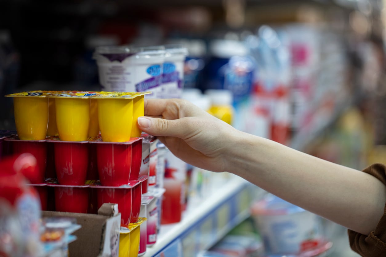 A person’s hand is reaching for a pack of yellow and red yogurt cups on a grocery store shelf, surrounded by various other overpriced products and dairy items.