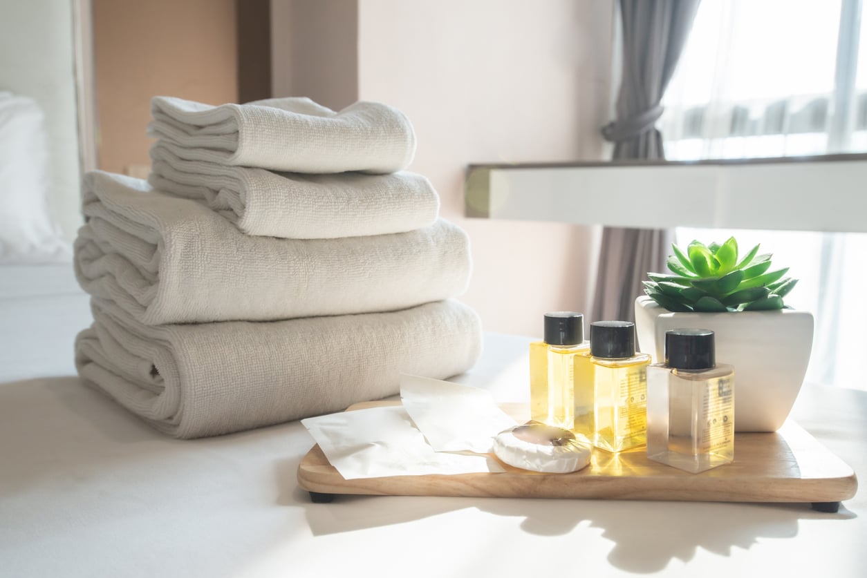 A stack of white towels, toiletry bottles, soap, and a small potted succulent plant arranged on a wooden tray evoke the comfort of top Las Vegas attractions, placed on a bed near a sunlit window with sheer curtains.