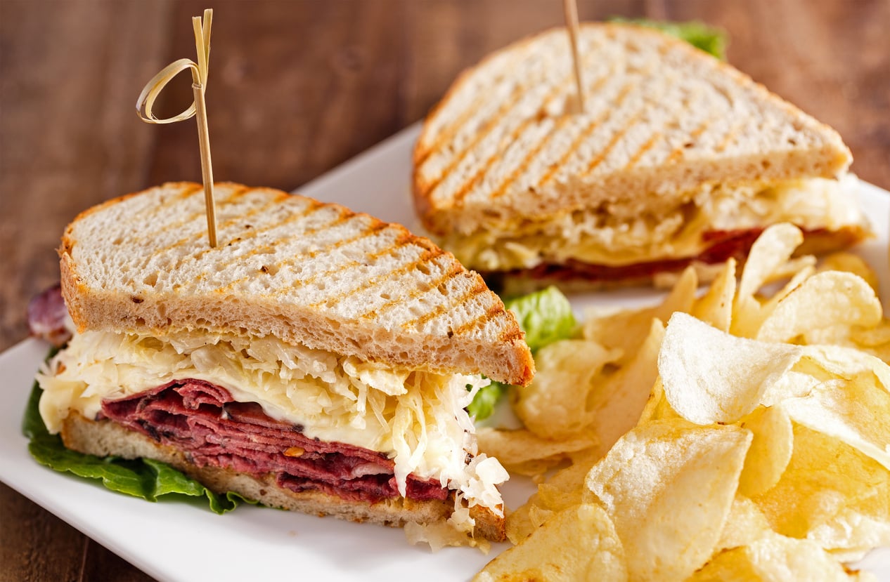 A close-up of a Reuben sandwich cut in half, filled with corned beef, sauerkraut, and cheese on grilled rye bread, served on a white plate with potato chips—proof that not all deliciousness comes from overpriced products.