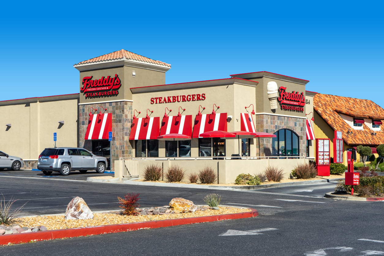 Freddy’s Steakburgers restaurant with red and white striped awnings, stone accents, and a parking lot with several cars under a clear blue sky.