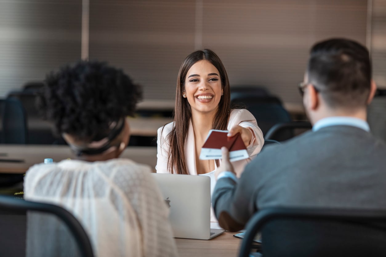 A smiling woman sitting at a desk hands a passport to a man across from her, while another woman sits beside him. They appear to be in a modern office, possibly sharing travel hacks before their next adventure.