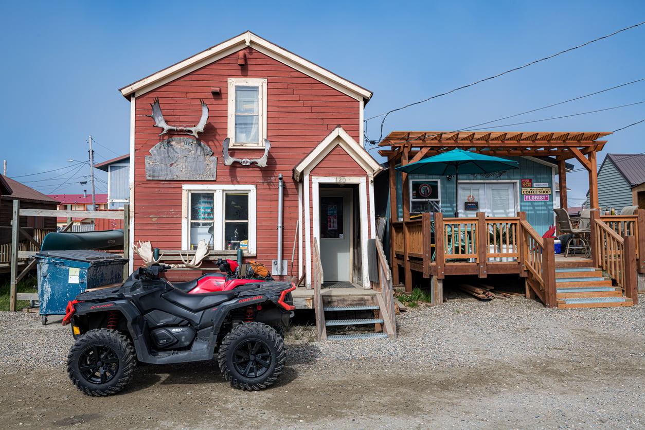 A red wooden building with antlers above the door stands next to a small cafe—an inviting spot typical of Small Business States—with a deck and teal awning. An ATV is parked out front on a gravel lot under a partly cloudy sky.