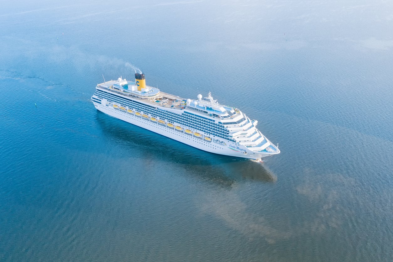A large white cruise ship sails through calm blue water, leaving a faint wake behind. Perfect for those seeking travel hacks, the ship features multiple decks with rows of windows and balconies, and a yellow funnel near the back.