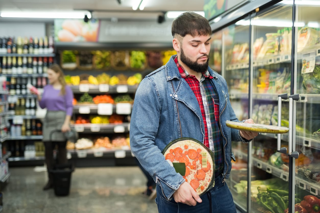 A man in a denim jacket holds a frozen pizza while opening a freezer door in a grocery store. Shelves of fruits, vegetables, and other overpriced products are visible, with a woman shopping in the background.